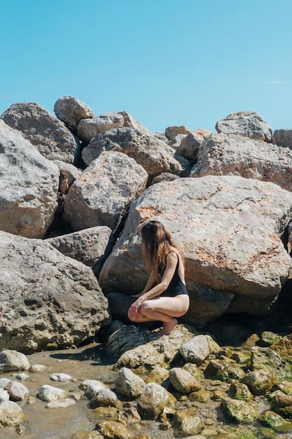 Woman in black swimsuit crouching on rock near sea
