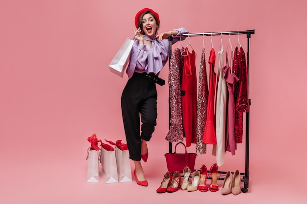 Woman in black trousers and purple blouse laughs, leaning on stand with elegant clothes on pink background.