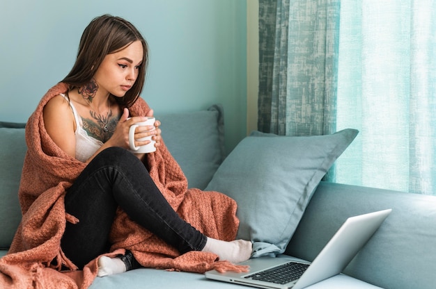 Donna in coperta che beve caffè a casa e lavora al computer portatile durante la pandemia