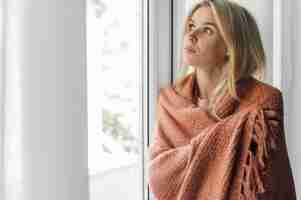 Free photo woman in blanket at home during the pandemic sitting next to window