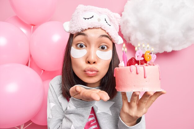 woman blows air kiss at camera keeps lips folded holds festive cake with burning candles prepares for celebrations applies beauty patches