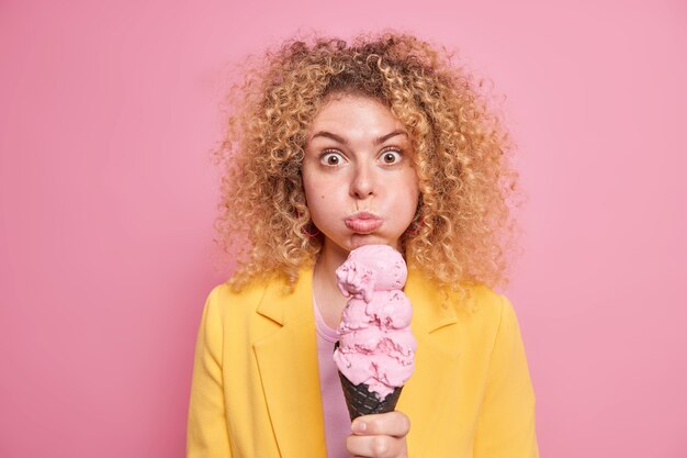 女性が頬を吹くと驚いた表情が美味しいアイスクリームを着てフォーマルなイエロージャケットを着てピンクの美味しい冷凍夏のデザートモデルを楽しんでいます