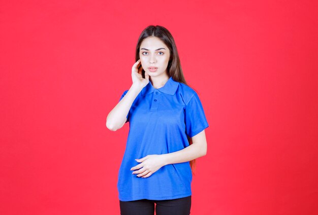 женщина в синей рубашке стоит на красной стене и выглядит смущенной и задумчивой.