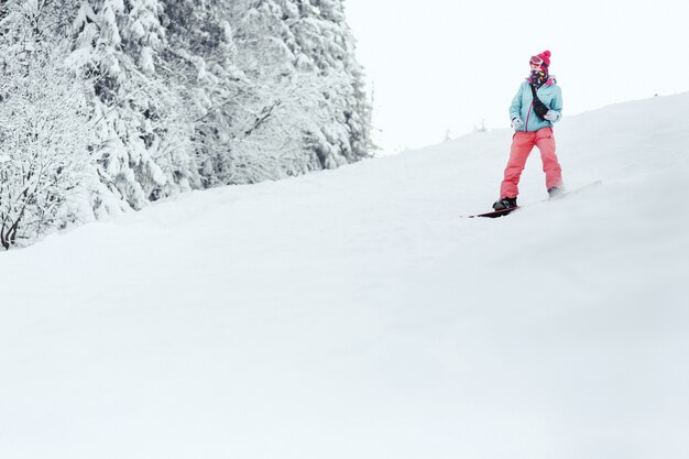 青いスキージャケットとピンクのズボンを着た女性が、スノーボードの雪が降った丘の下を行く