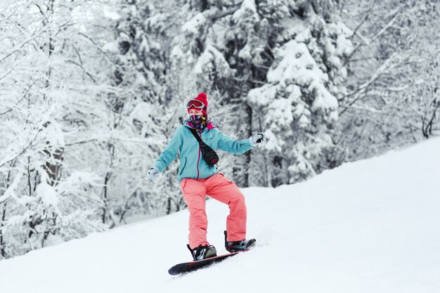 Woman in blue ski jacket and pink pants stands on the snowboard somewhere in winter forest 