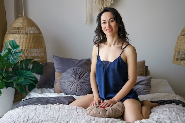 A woman in a blue slip sits on the bed and smiles. The concept of rest on a day off.