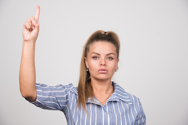 Woman in blue sweater pointing at up on gray wall.