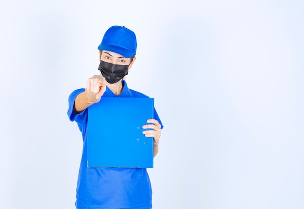 Woman in blue uniform and black face mask holding a blue folder and pointing at someone aside. 