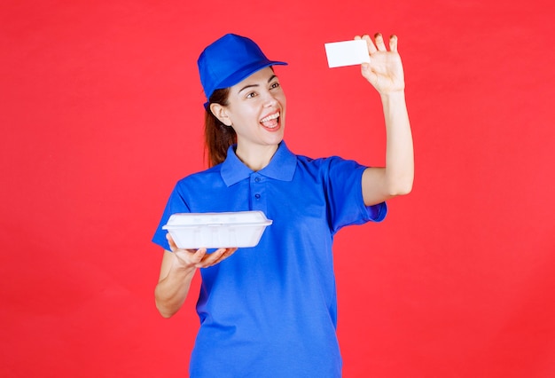 Женщина в синей форме держит белую пластиковую коробку для еды на вынос для доставки и представляет свою визитную карточку клиенту.