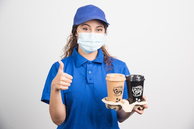 Woman in blue uniform with medical mask holding two cups of coffee and shows ok gesture on white