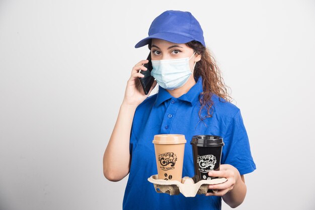 Woman in blue uniform with medical mask speaking on phone and holding two cups of coffee on white