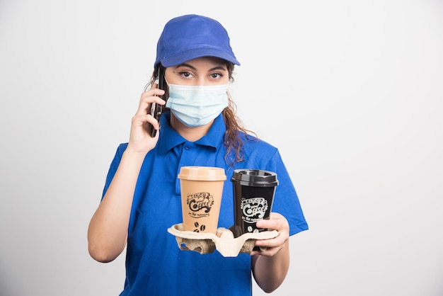 Женщина в синей форме с медицинской маской разговаривает по телефону и держит две чашки кофе на белом
