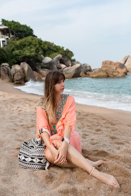 Женщина в летнем платье Бохо, сидя на песке у моря. Тропическое настроение.