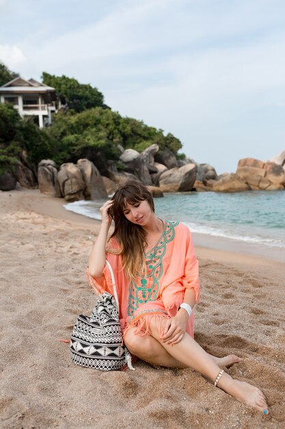 Woman in boho summer dress sitting on sand near sea. Tropical mood.