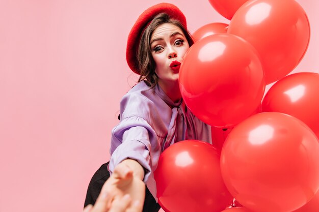 Woman in bright beret and purple blouse is looking at in surprise, reaching for camera and holding balloons.