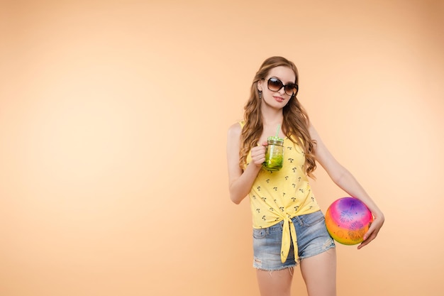 Free photo a woman in bright clothes a yellow tshirt and denim shorts sunglasses holds a ball and cold lemonade in her hands on an isolated background