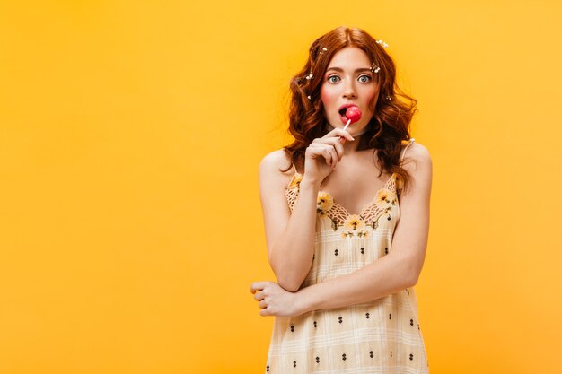 Woman in bright summer dress eats lollipop. Curly woman with wild flowers in her hair in surprise poses on orange background.