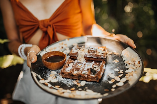 Woman in brown bra holds plate with waffles, chocolate sauce and peanuts