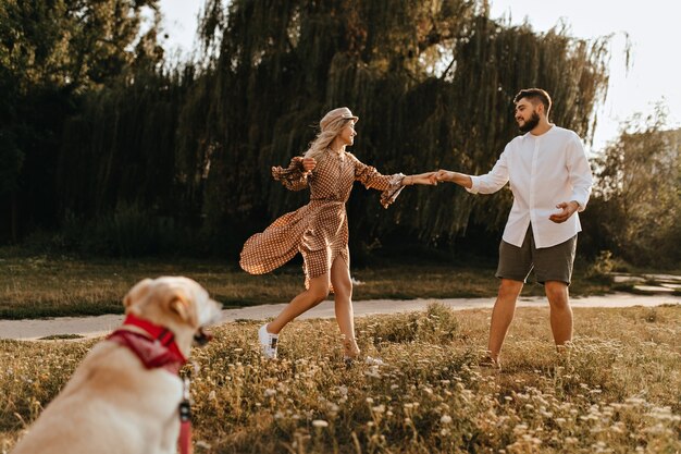 茶色のドレスと帽子をかぶった女性とショートパンツとシャツを着た男性が犬と一緒に歩いて公園を騙している。