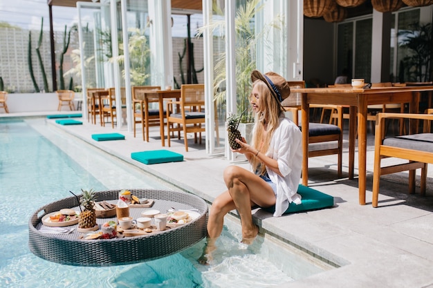 woman in brown hat drinking pineapple cocktail while resting near pool. Magnificent blonde female model chilling at resort cafe in weekend morning.