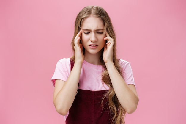 Woman cannot focus during test having answer slipped away touching temples looking down concerned and troubled having headache or problems with memory standing displeased over pink wall.