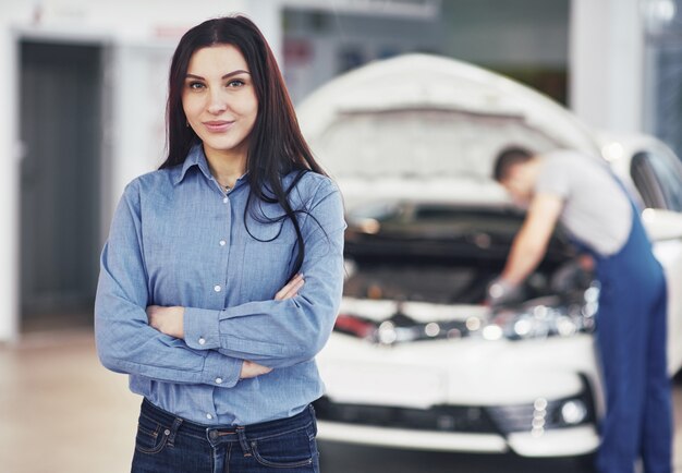 機械サービスを取得する車のガレージで女性。メカニックは車のボンネットの下で動作します