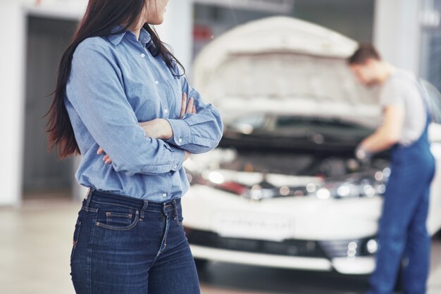 機械サービスを取得する車のガレージで女性。メカニックは車のボンネットの下で動作します