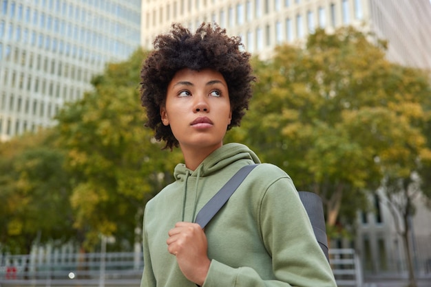 woman carries yoga mat rests after training concentrated into distance wears sweatshirt poses. Healthy lifestyle and wellbeing concept