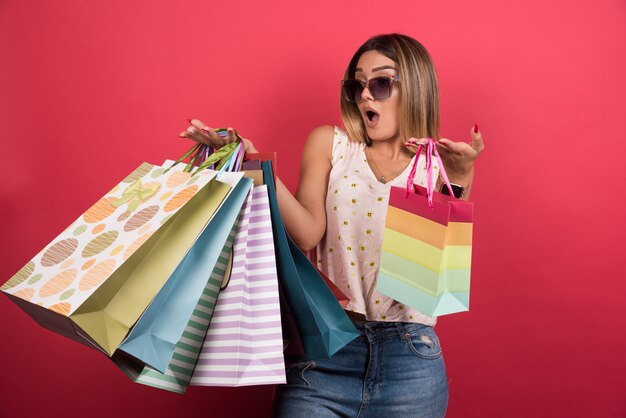 Woman carrying shopping bags with surprised expression on red wall . 