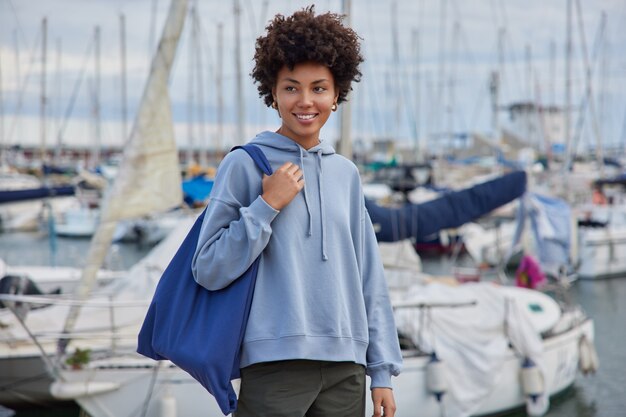 woman in casual clothes carries fabric bag strolls in harbor on pier breathes fresh marine air waits for boat looks happily away has travel at sea