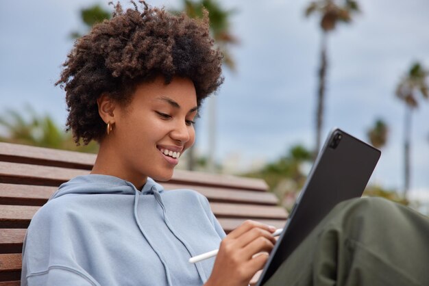 woman in casual clothes draws pictures on modern tablet feels glad sits on wooden bench spends free time on hobby enjoys good sunny weather works freelance uses modern technology