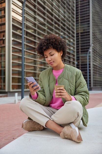 カジュアルな服装の女性が足を組んで座っている携帯電話を使用してオンライン投稿情報をソーシャルメディアで飲む使い捨てカップのポーズから芳香性のコーヒーを飲む