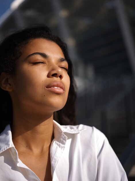 woman in casual white shirt touching her hair while posing with eyes closed outdoors on a warm sunny day. Selective focus