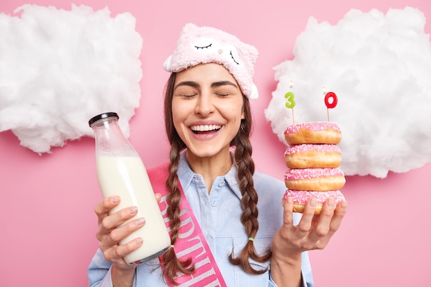 woman celebrates 30th anniversary keeps eyes closed and laughs poses with stack of delicious donuts and bottle of milk wears sleepmask shirt expresses happiness