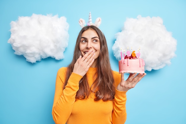 woman celebrates birthday holds strawberry cake cons mouth and chuckles dressed in casual jumper isolated on blue 