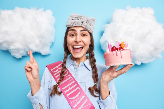 woman celebrates birthday makes wish before blowing candles on festive cake keeps fingers crossed wears sleepmask and shirt isolated on blue 