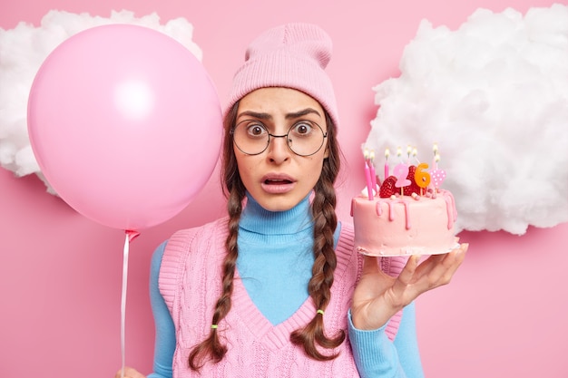 woman celebrates her 26th birthday holds cake with burning candles and inflated balloon wears pink hat turtleneck big round spectacles stands indoor