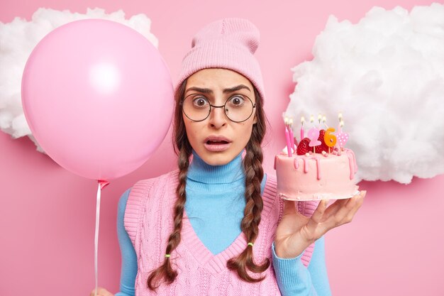 26 歳の誕生日を祝う女性は、ろうそくを燃やしてケーキを持ち、膨らませた風船はピンクの帽子のタートルネックを着て、大きな丸い眼鏡を屋内に立てます