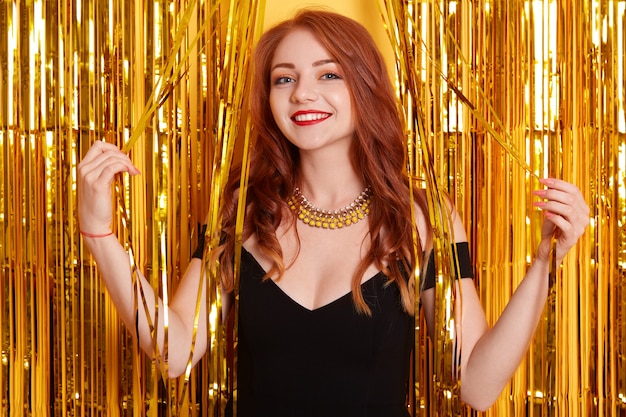 Woman celebrating New Year party, beautiful smiling girl in black dress over golden tinsel on space, 