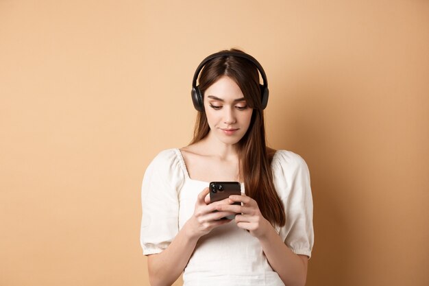 베이지색 배경에 서 있는 무선 이어폰으로 휴대폰으로 채팅하고 음악을 듣는 여성