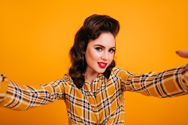 Free photo woman in checkered shirt taking selfie. studio shot of smiling pinup girl posing on yellow background.