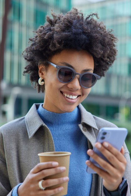 女性はスマートフォンで通知をチェックしますニュースを読みますオンラインドリンク持ち帰りコーヒー笑顔は喜んでサングラスをかけ、街の中心部でコートのポーズはモダンなガジェットを使用しています