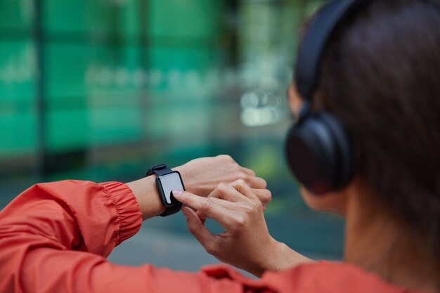  woman checks results of fitness training on smartwatch listens music via headphones dressed in anorak poses on blurred 