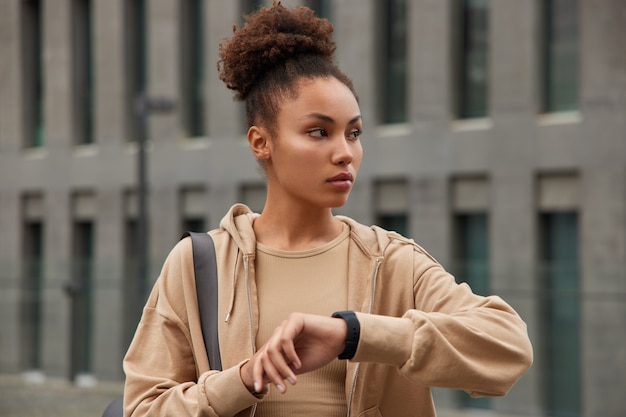 woman checks time on digital smartwatch tracks her wotkout performance looks away dressed in comfortable sportswear poses against city building
