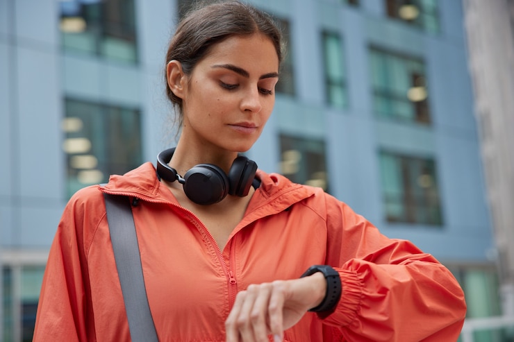 Free Photo woman checks time on smartwatch uses headphones for listening audio track dressed in jacket waits for coach poses