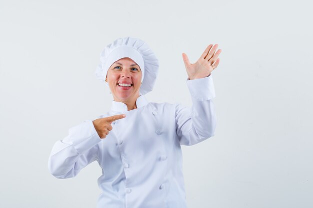 женщина-шеф-повар позирует, как указывая на ее руку, держащую телефон в белой форме, и выглядит сосредоточенной.
