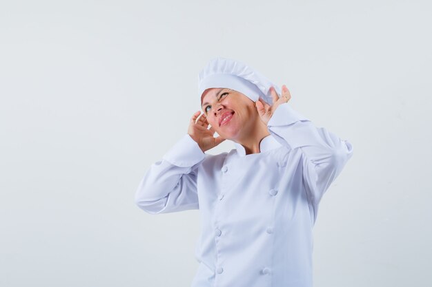 흰색 제복을 입은 여자 요리사는 이어폰을 착용하고 즐겁게 보이는 것처럼 포즈를 취합니다.