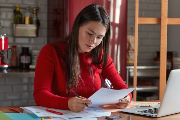 Woman chief architect works with documentation, confirms project, holds paper and glasses in hands, poses at kitchen, chats with marketing experts on laptop computer. People and career concept