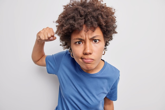 woman clenches fist and looks irritated frowns face doesnt like somethning wears casual blue t shirt isolated on white. Irritated female model feels rage