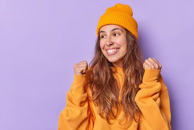 женщина сжимает кулаки празднует достижения, носит повседневный оранжевый джемпер и шляпу радуется успеху, изолирована на фиолетовом фоне с пустым пространством для копии слева
